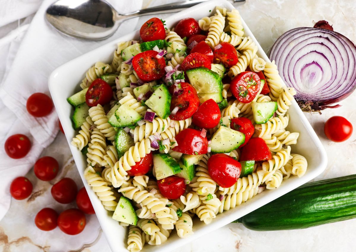 A bowl of rotini pasta salad with cherry tomatoes, cucumber, and chopped red onion, placed on a marble surface with a spoon, tomatoes, and half a red onion nearby.