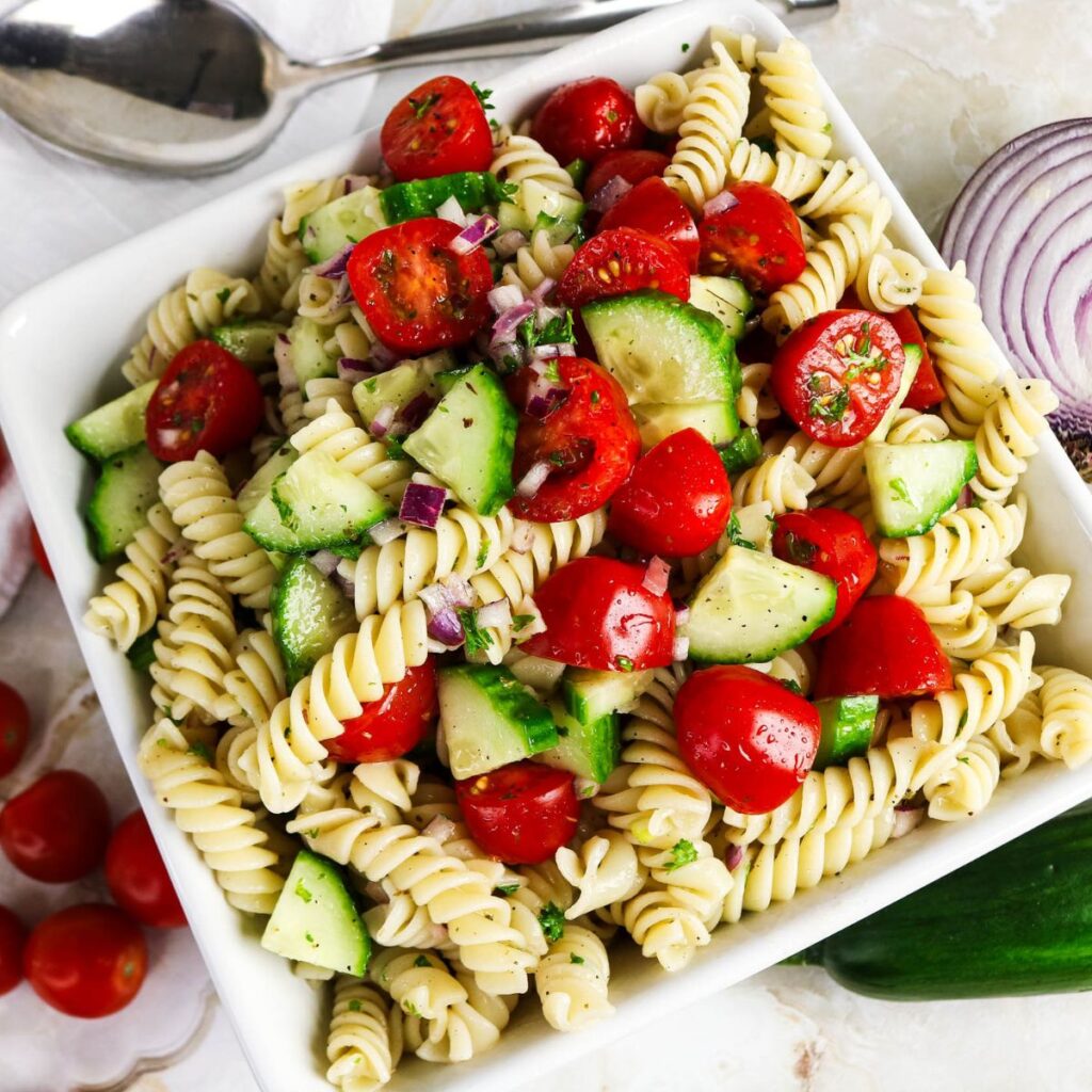 A square white dish filled with rotini pasta salad mixed with cherry tomatoes, cucumber slices, chopped red onion, and herbs. Spoons and vegetables are on the side.