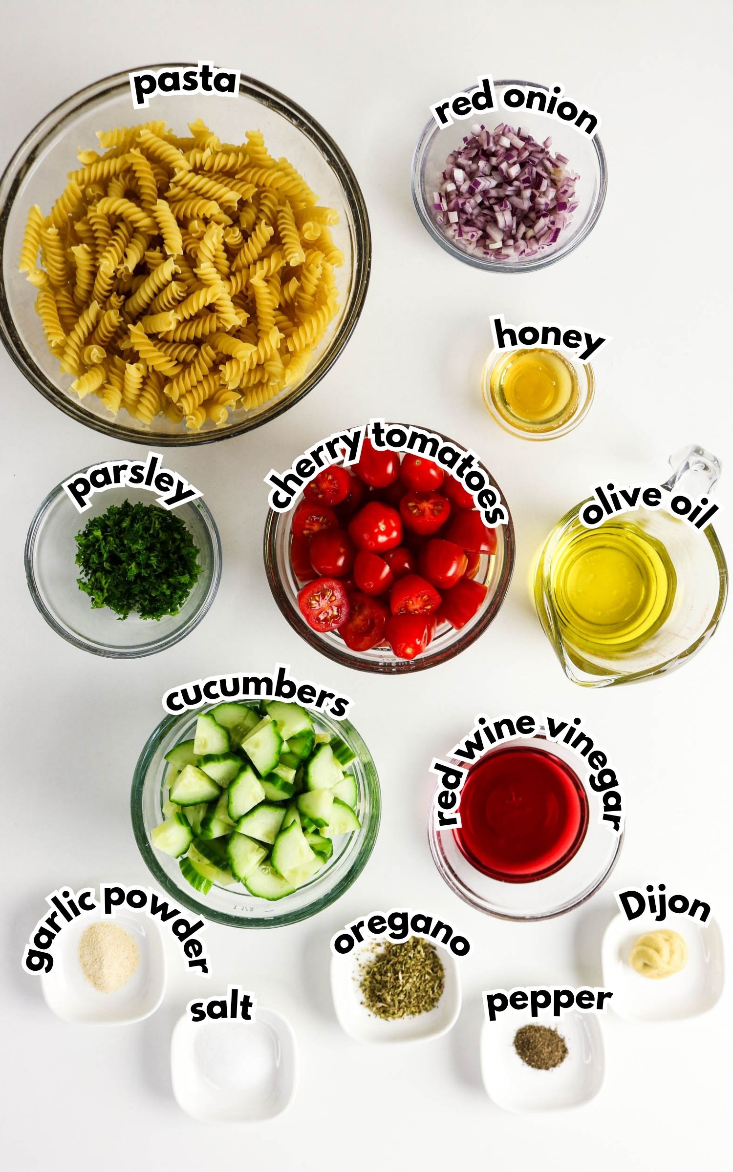 Ingredients for a pasta salad are laid out on a white surface, including pasta, cherry tomatoes, cucumbers, red onion, parsley, olive oil, red wine vinegar, and various seasonings.