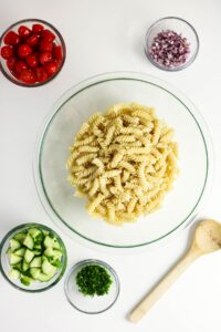 A glass bowl of cooked rotini pasta is surrounded by bowls of chopped tomatoes, red onion, cucumber, parsley, and a wooden spoon on a white surface.