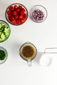 Bowls of chopped cucumber, cherry tomatoes, red onion, parsley, and a measuring cup of salad dressing with a whisk, arranged on a white surface.
