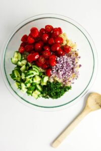 A glass bowl with uncooked pasta, halved cherry tomatoes, diced cucumber, chopped red onion, and parsley. A wooden spoon lies beside the bowl on a white surface.