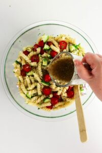 A hand pours vinaigrette dressing onto a bowl of pasta salad with rotini, cherry tomatoes, cucumbers, and red onions.
