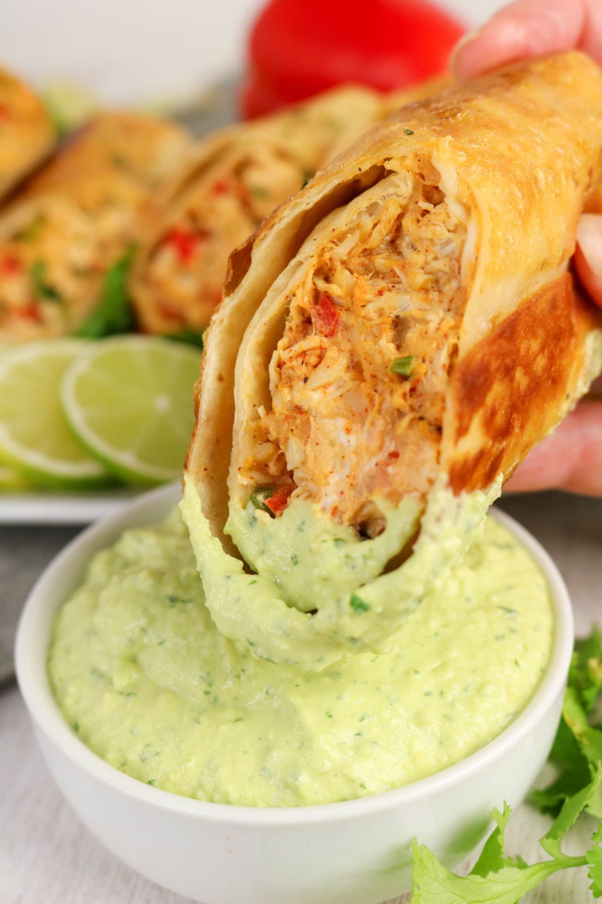 A hand dips a crispy rolled tortilla filled with chicken and vegetables into a bowl of creamy green dip, with lime slices and more tortillas in the background.