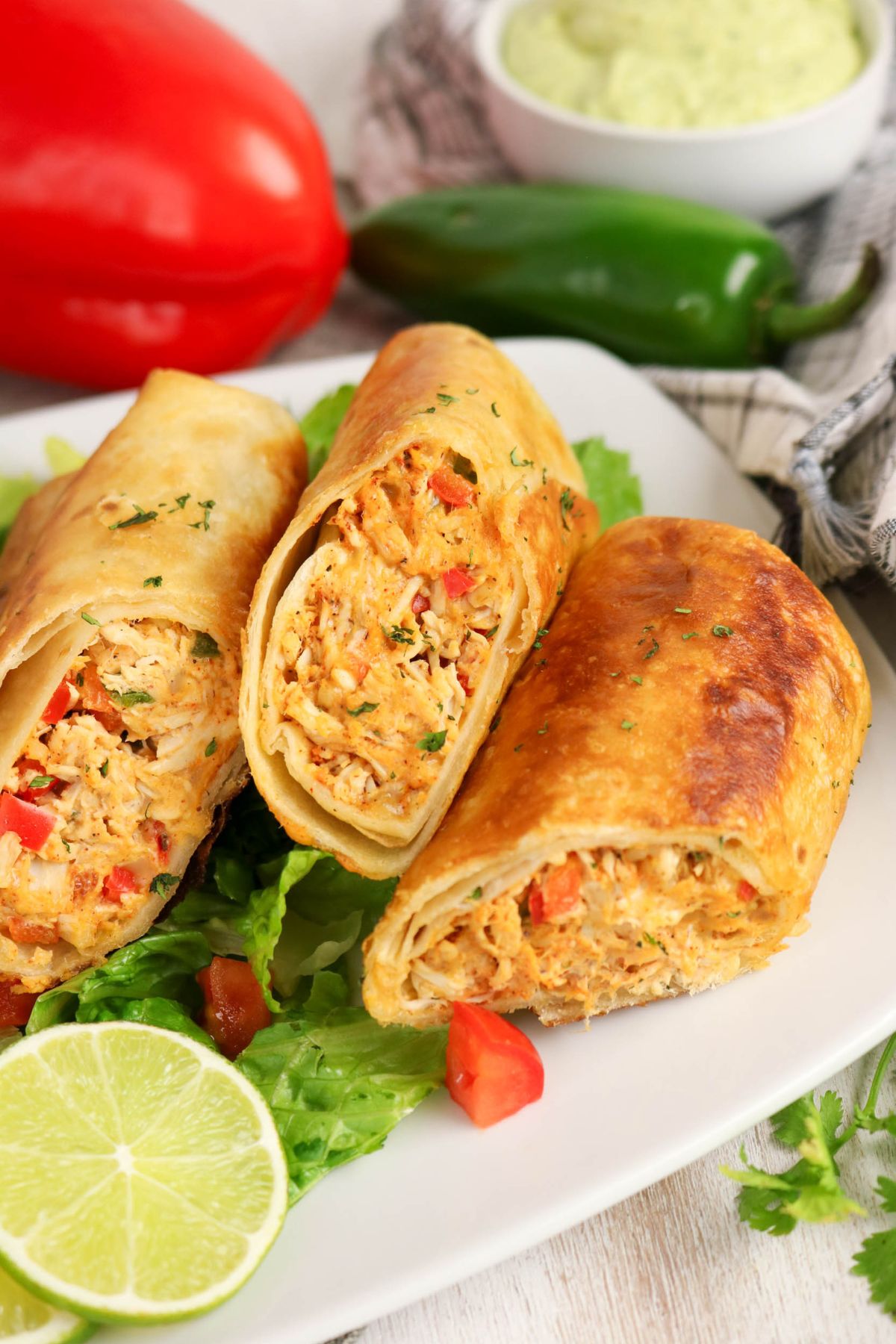 Three crispy chimichangas filled with shredded chicken and vegetables are served on lettuce with diced tomatoes and a lime wedge, with a red pepper, jalapeño, and dip in the background.