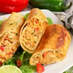 Three crispy baked chimichangas filled with shredded chicken and diced vegetables are served on a bed of lettuce, with a lime wedge and sliced peppers in the background.