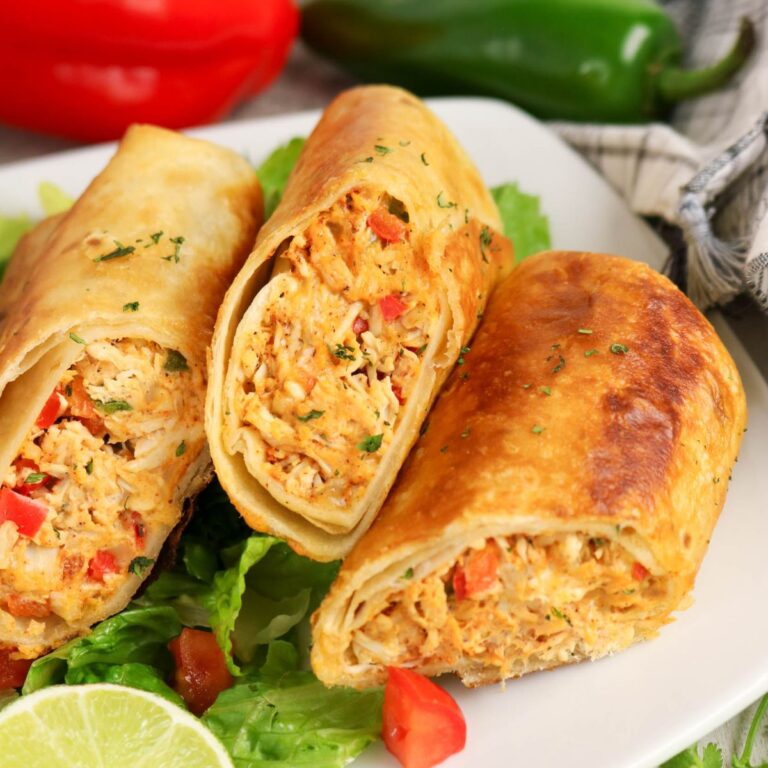 Three crispy baked chimichangas filled with shredded chicken and diced vegetables are served on a bed of lettuce, with a lime wedge and sliced peppers in the background.