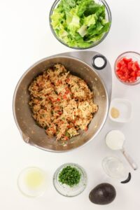 Overhead view of a mixing bowl with shredded chicken and seasonings, surrounded by bowls of lettuce, diced tomato, chopped herbs, avocado, and other ingredients on a white surface.