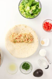 A flour tortilla with a chicken mixture is on a white surface, surrounded by bowls of lettuce, diced tomatoes, seasonings, lime juice, cilantro, an avocado, and sour cream.
