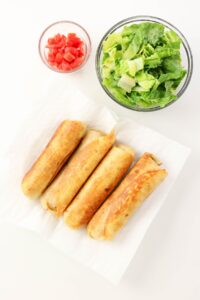 Five browned taquitos on a paper towel with a bowl of chopped lettuce and a bowl of diced tomatoes on a white surface.