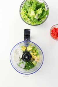 A food processor with avocado, yogurt, and herbs, next to bowls of chopped lettuce and diced tomatoes on a white surface.