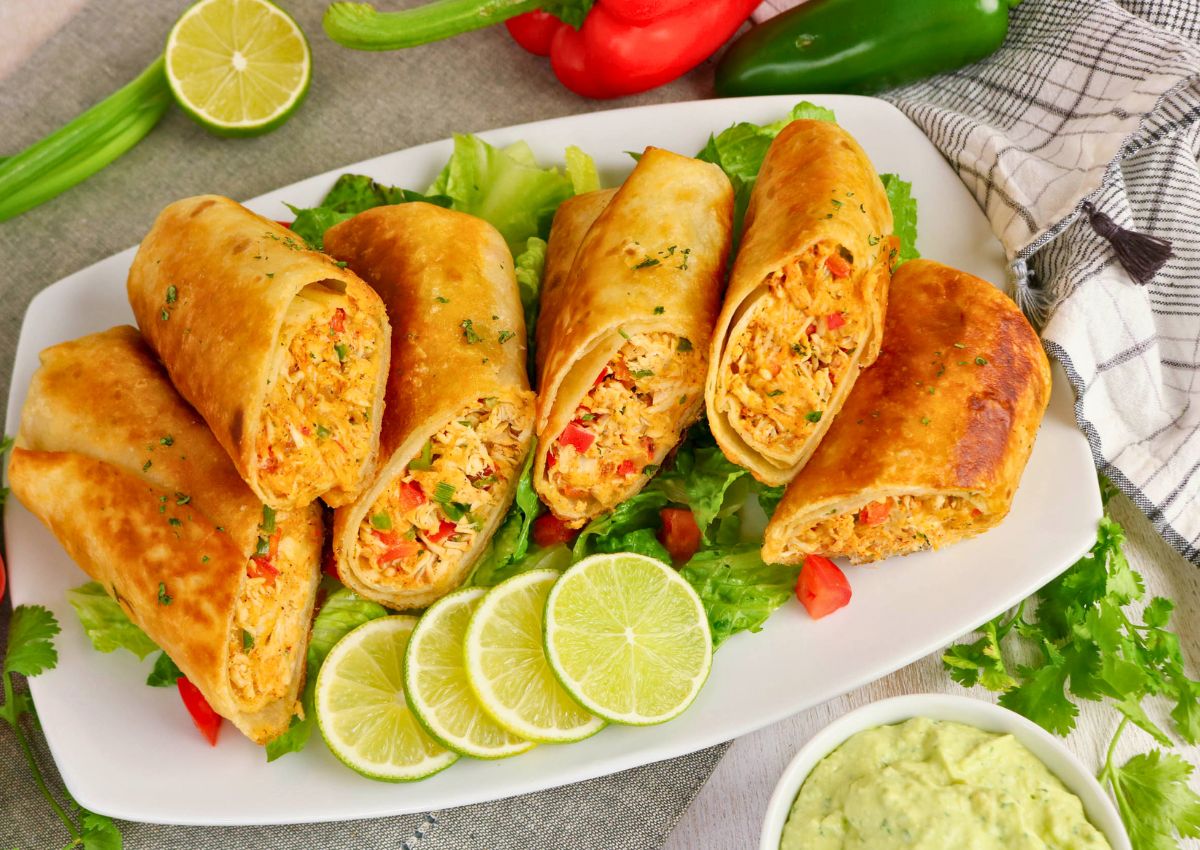 A plate of five cut chicken chimichangas filled with shredded chicken and vegetables, served on lettuce with lime slices, avocado dip, and assorted peppers nearby.