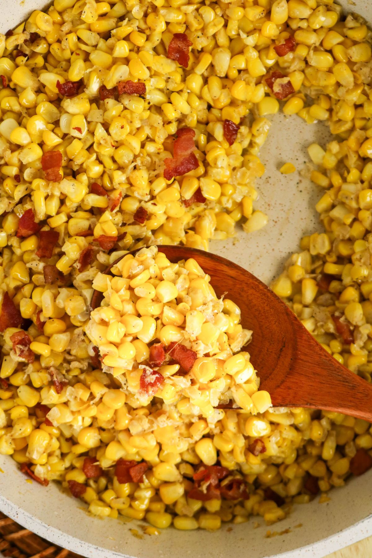 Close-up of a pan filled with cooked corn and bacon pieces, with a wooden spoon scooping some of the mixture.