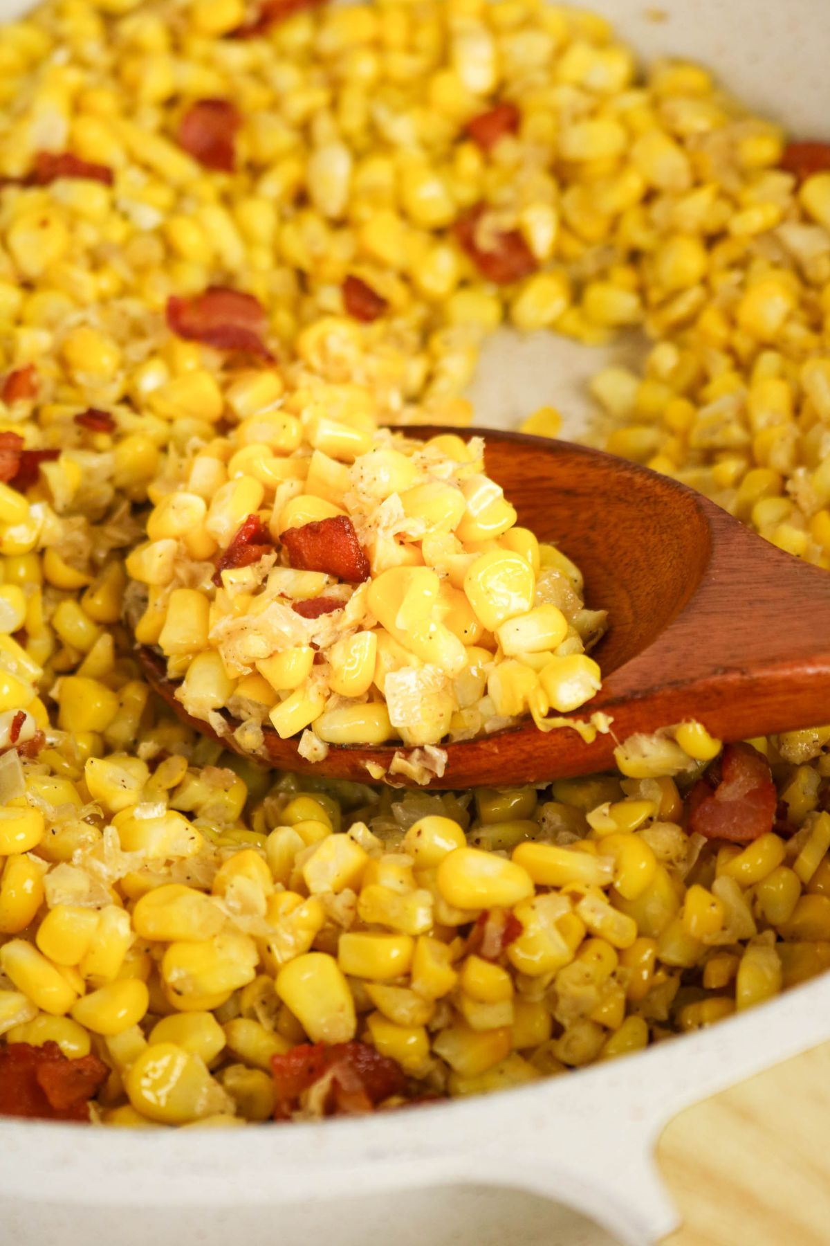 A wooden spoon scoops cooked corn mixed with diced bacon and onions from a white pan.