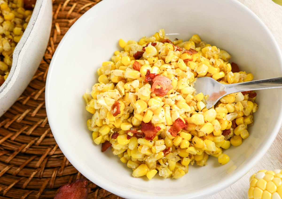 A white bowl filled with creamed corn mixed with bacon pieces, with a fork resting inside; the bowl sits on a woven placemat.