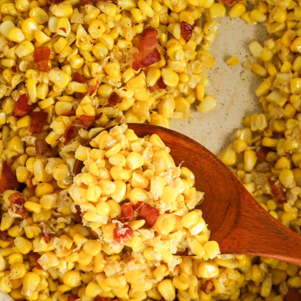 Close-up of creamy corn and bacon mixture in a pan, with a wooden spoon scooping some of the dish.