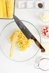 Overhead view of a glass bowl with cut corn kernels and a corn cob, a knife resting on the bowl, and various ingredients like butter, bacon, and seasonings arranged around it.