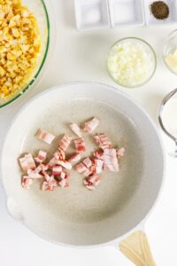 Uncooked bacon pieces in a white skillet with chopped onions, corn, butter, and seasonings arranged nearby on a white surface.