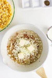 A white skillet with chopped onions, butter, and seasoning, next to a bowl of corn and a measuring cup of milk on a white surface.