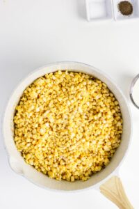 A white skillet filled with cooked corn kernels sits on a white surface, with a wooden spoon handle visible and small dishes of salt and pepper nearby.