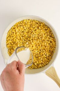 A hand pours milk from a measuring cup into a skillet filled with cooked corn kernels.