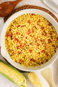 A white skillet filled with cooked corn kernels and bits of bacon sits on a woven mat next to a wooden spoon and fresh ears of corn.