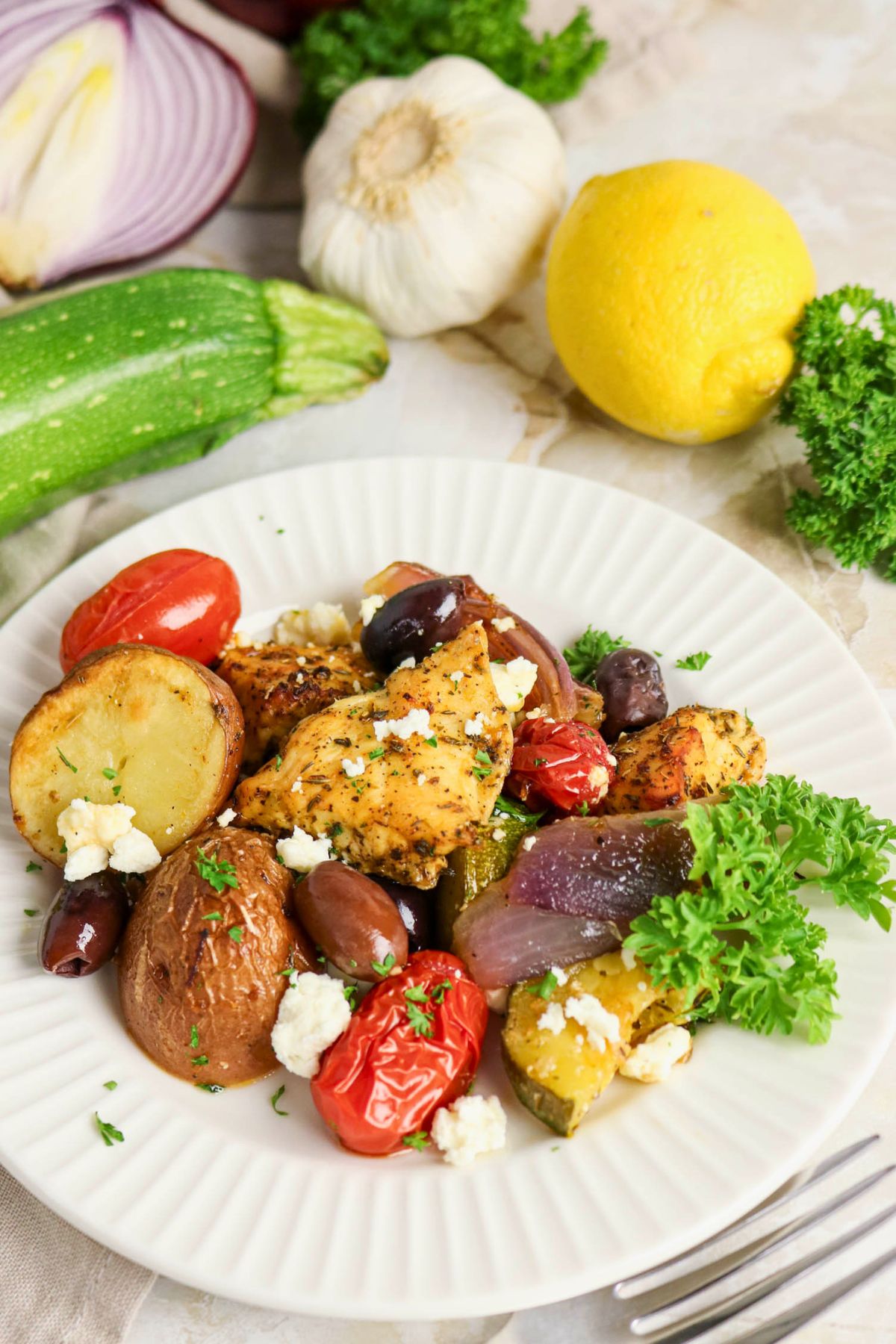 A plate of roasted chicken, potatoes, tomatoes, olives, red onion, zucchini, and feta cheese garnished with parsley. Lemon, garlic, zucchini, and parsley are in the background.