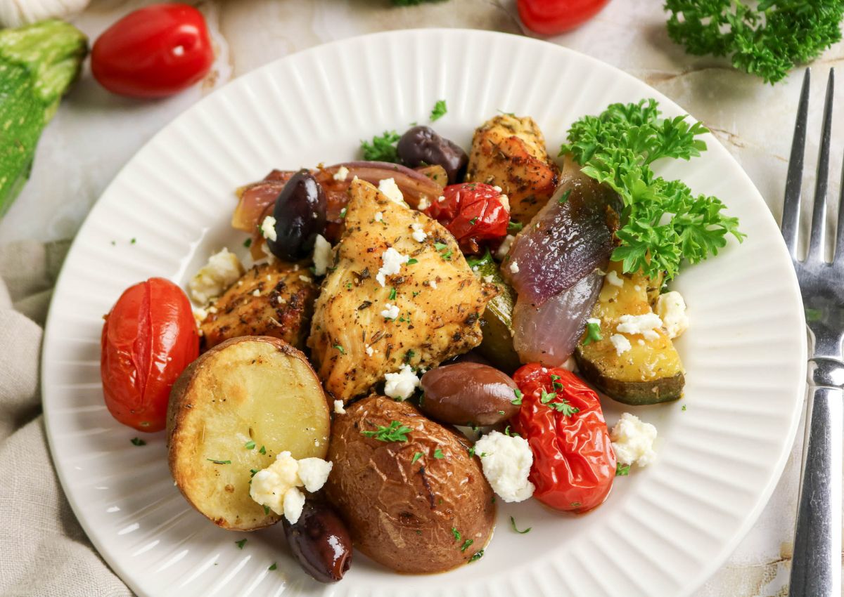 A plate of roasted chicken, potatoes, cherry tomatoes, red onion, zucchini, olives, and crumbled feta, garnished with parsley.