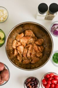 Raw, seasoned chicken pieces in a metal bowl surrounded by bowls of chopped vegetables, olives, cherry tomatoes, cheese, herbs, and containers of salt and pepper on a white surface.