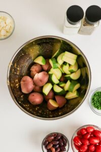 A metal bowl with chopped red potatoes and zucchini, surrounded by small bowls of feta cheese, olives, cherry tomatoes, herbs, and salt and pepper shakers on a white surface.