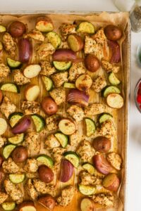 Sheet pan with roasted chicken chunks, red potatoes, zucchini slices, and red onion on parchment paper.