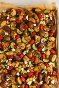 Sheet pan with roasted chicken, red potatoes, zucchini, red onions, cherry tomatoes, olives, and crumbled feta cheese on parchment paper.