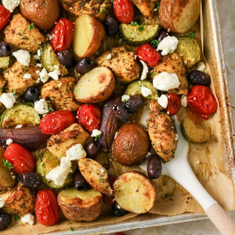 Sheet pan of roasted chicken, potatoes, zucchini, grape tomatoes, red onions, black olives, and crumbled feta cheese on parchment paper with a white serving spoon.
