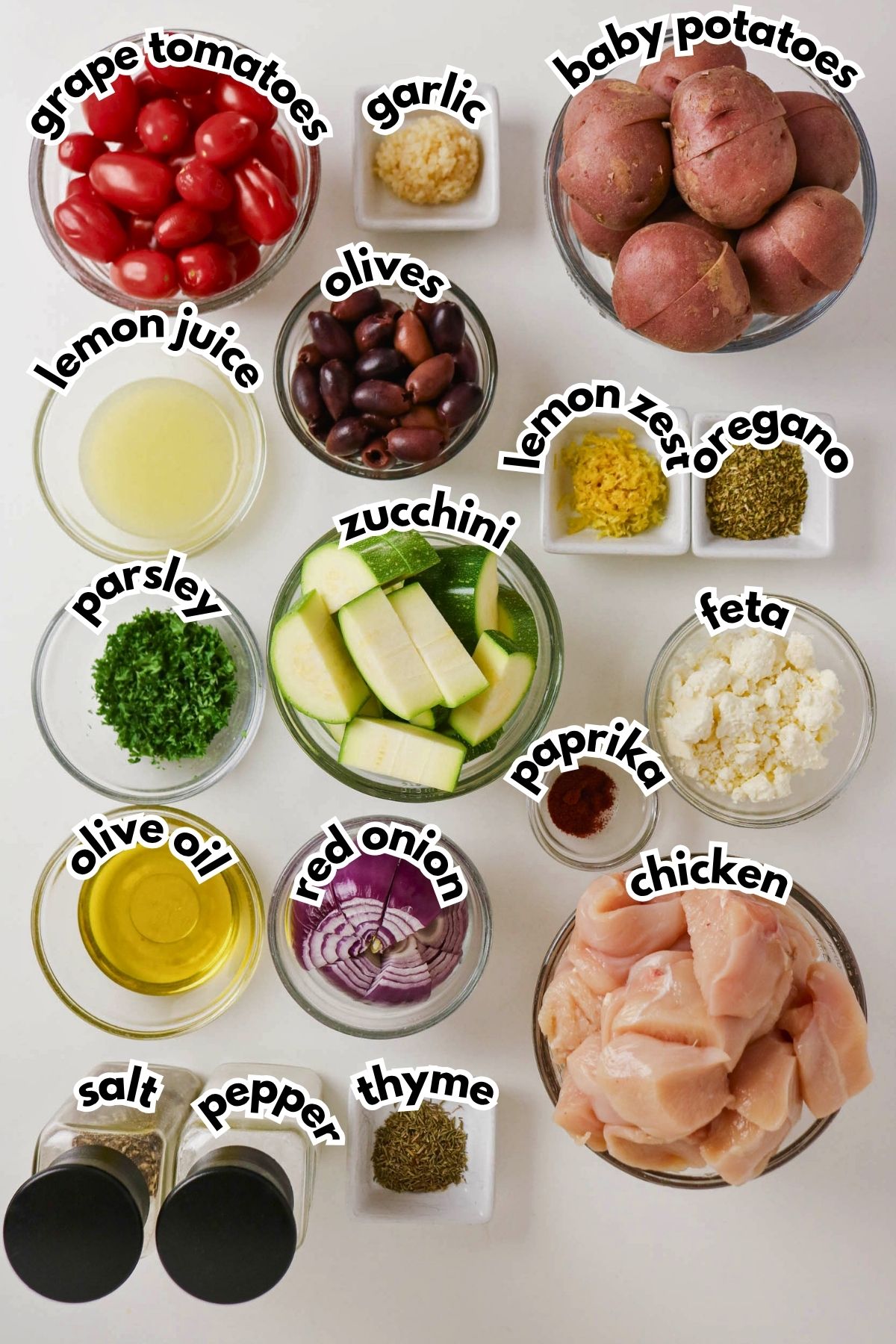 Ingredients for a recipe arranged on a white surface, including chicken, grape tomatoes, baby potatoes, zucchini, olives, feta, onion, lemon juice, spices, olive oil, and herbs.