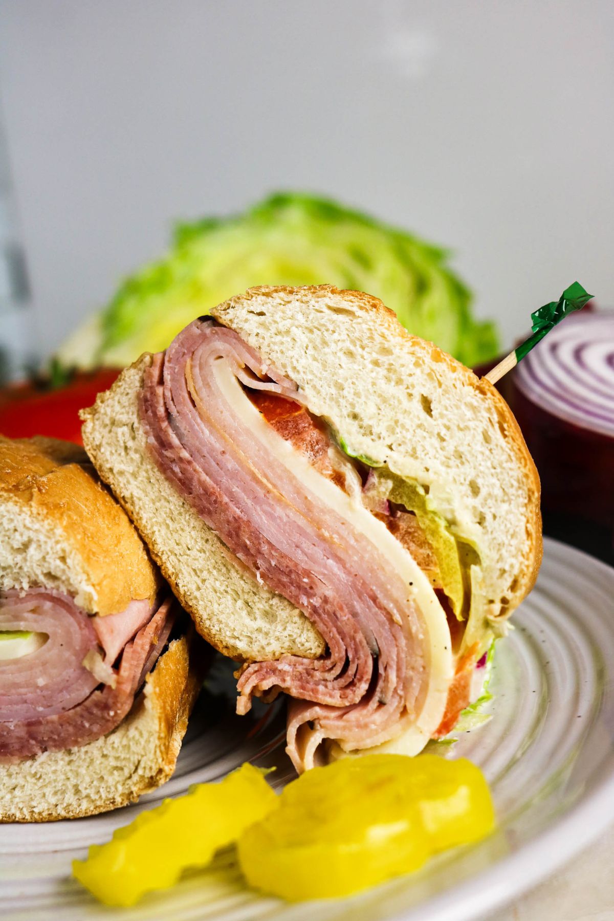 A sandwich with layers of deli meats, cheese, lettuce, and tomato is cut in half on a plate, with pickle slices in the foreground and lettuce and onion in the background.