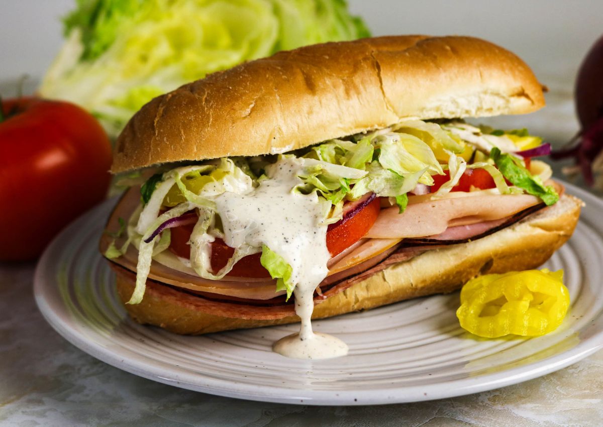 A sandwich with sliced deli meat, tomato, lettuce, red onion, and creamy dressing on a hoagie roll, served on a plate with a pepper slice.