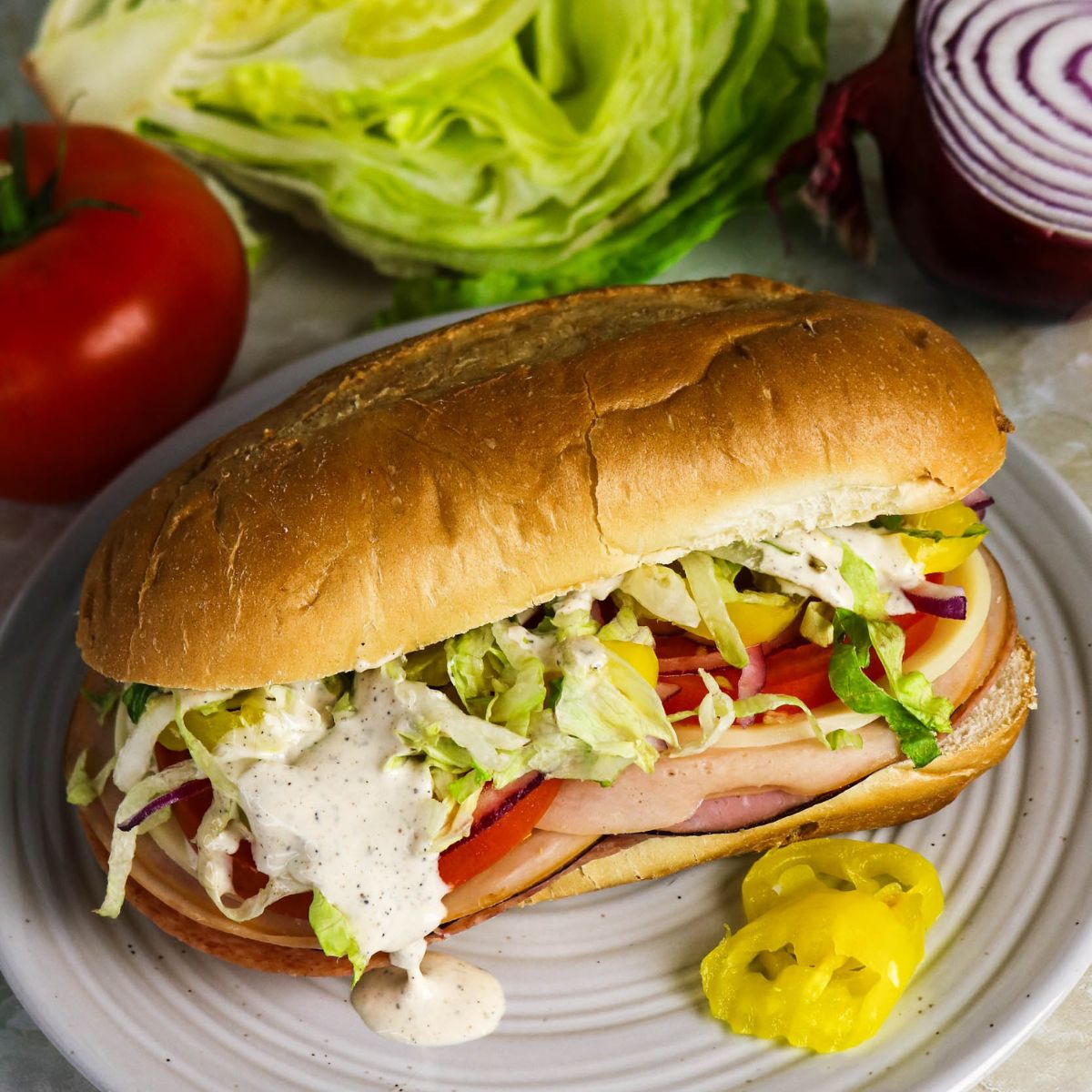 A sub sandwich with deli meat, lettuce, tomato, red onion, and creamy dressing on a plate, garnished with banana peppers. Lettuce, tomato, and onion are in the background.
