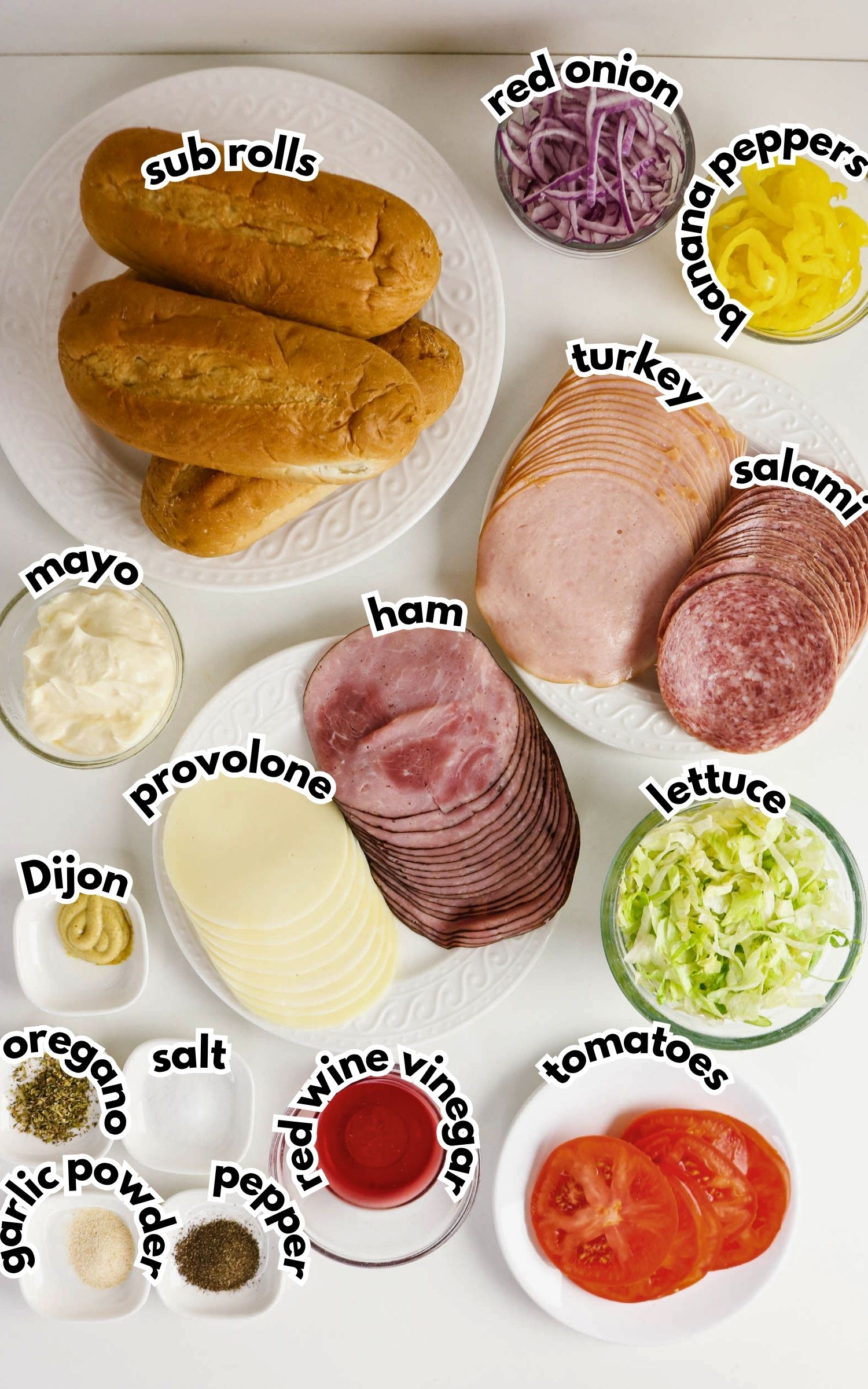Assorted sandwich ingredients on plates and bowls, including sub rolls, cold cuts, cheese, vegetables, and seasonings, all labeled with text.