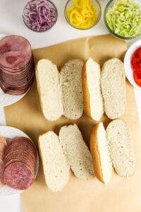 Four sliced hoagie rolls on parchment paper surrounded by plates of deli meats, sliced red onions, banana peppers, shredded lettuce, and sliced tomatoes.