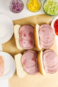 Four sandwich rolls filled with slices of ham are on parchment paper, surrounded by plates and bowls of sliced vegetables, including onions, peppers, lettuce, and tomatoes.