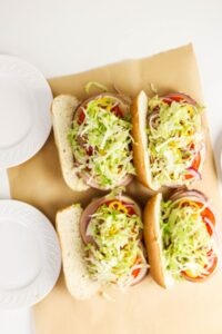 Four sub sandwiches with sliced deli meat, tomato, red onion, and shredded lettuce on a sheet of brown parchment paper, next to two stacked white plates.