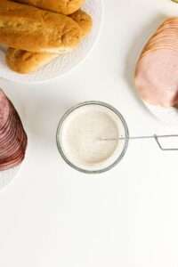 A bowl of creamy white sauce with a whisk, surrounded by plates of sliced ham, turkey, and sandwich rolls on a white surface.