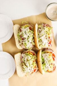 Four sub sandwiches topped with shredded lettuce, sliced onions, tomatoes, and creamy dressing, arranged on parchment paper beside empty white plates and a jar of extra dressing.