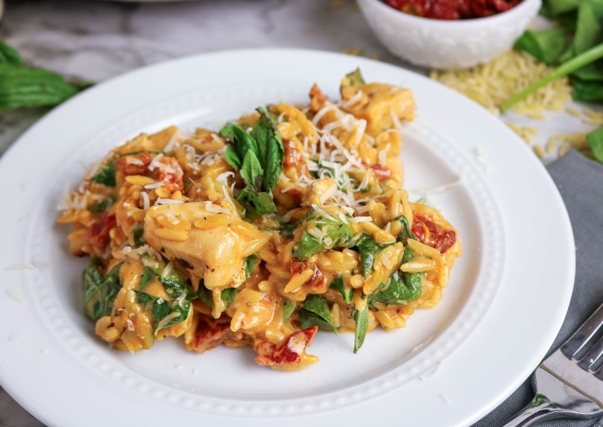 A white plate with creamy pasta mixed with chicken, spinach, sun-dried tomatoes, and shredded cheese, garnished with fresh basil.