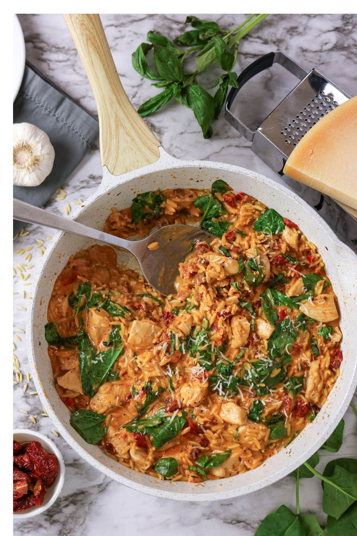 A skillet filled with creamy orzo, spinach, sun-dried tomatoes, and chicken, garnished with shredded cheese. Fresh basil, garlic, and a block of cheese are nearby on the marble surface.