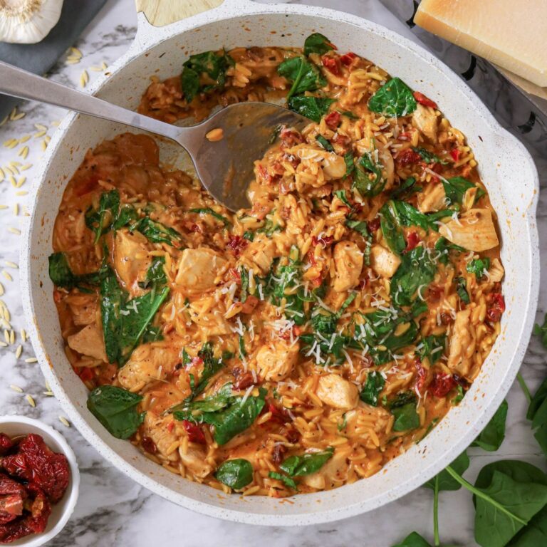 A skillet filled with creamy orzo pasta, pieces of chicken, spinach, sun-dried tomatoes, and grated cheese, with a serving spoon inside.