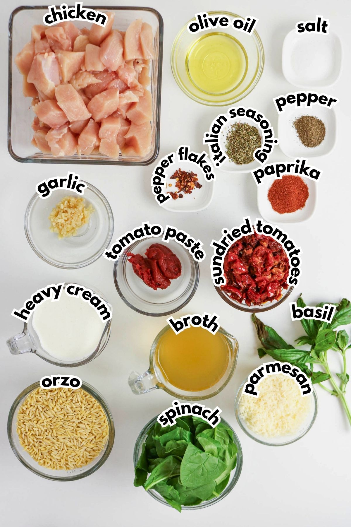 Ingredients for a recipe displayed on a white surface, including chicken, olive oil, spices, garlic, tomato paste, sundried tomatoes, basil, orzo, broth, spinach, parmesan, and heavy cream.