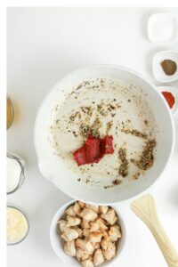 A white skillet with spices and tomato paste, surrounded by small bowls of cooked chicken, grated cheese, cream, broth, and seasonings on a white surface.