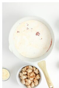A white skillet with cream and seasoning, next to a bowl of cooked chicken pieces and a small bowl of grated cheese on a white background.