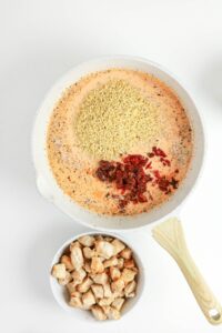 A pan with uncooked orzo, cream sauce, spices, and sun-dried tomatoes sits beside a bowl of cooked, seasoned chicken pieces on a white surface.